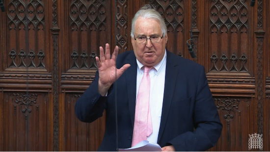 The Viscount Stansgate addressing the House of Lords, with a hand raised and wearing a suit with pink tie